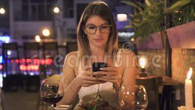 优雅的女人用<strong>手机</strong>坐在晚上的餐厅的餐桌上。 漂亮的女人<strong>看</strong>着智能<strong>手机</strong>